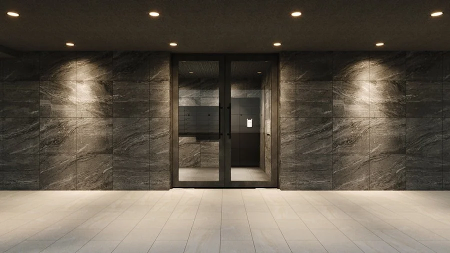 モダンな内装のエントランスまたは廊下空間です。壁は石目調のダークなタイルで覆われ、床は明るい色のタイル張りです。中央にはガラスの二重扉があり、天井のダウンライトが壁面と床を照らし、洗練された高級感を演出しています。