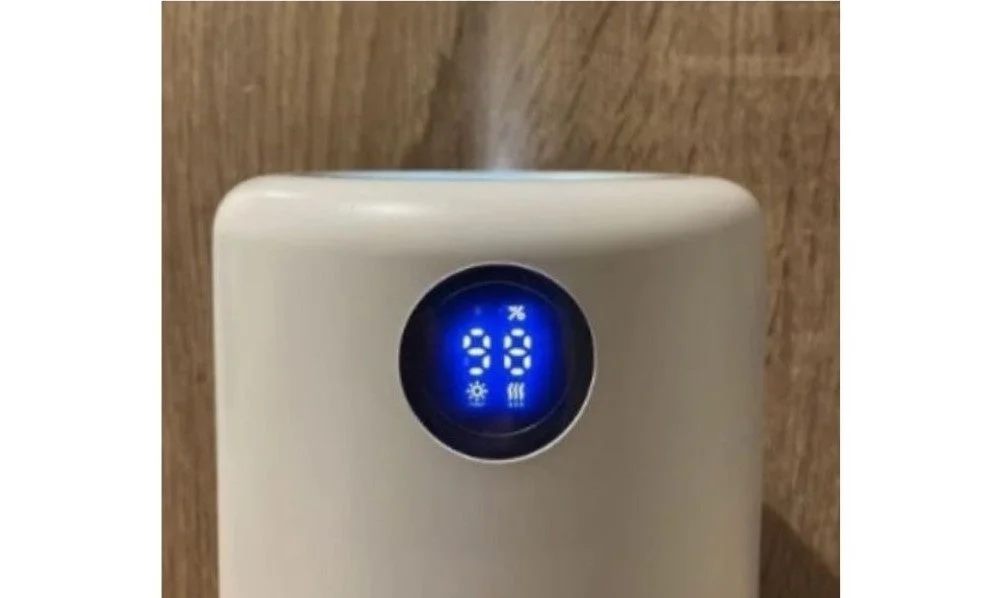 白い加湿器またはアロマディフューザーがミストを噴出しており、青色のデジタルディスプレイには「98%」と表示されています。木目調の壁を背景に、動作中の様子が捉えられています。