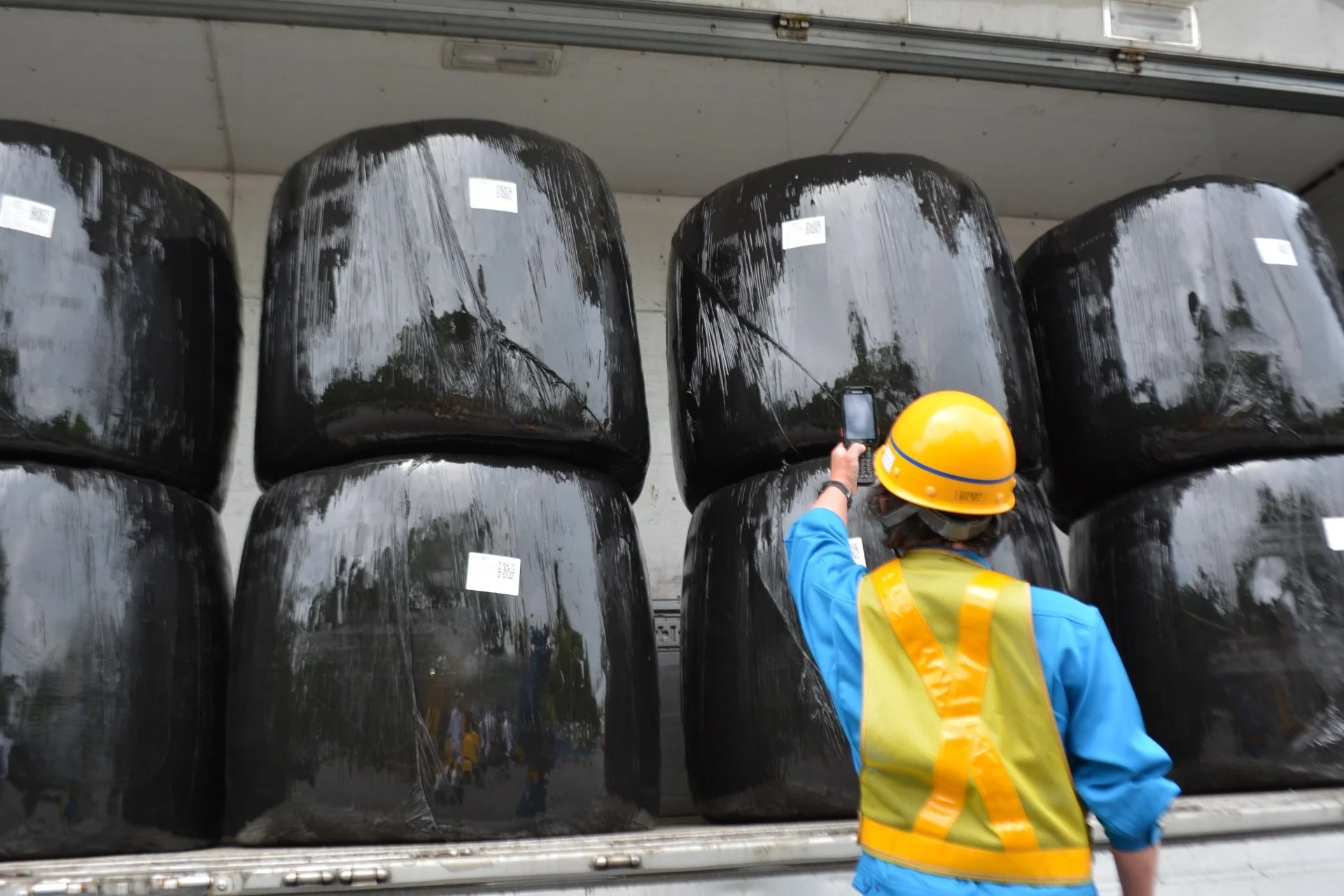 トラックの荷台に積まれた黒い牧草ロールを、安全ヘルメットとベストを着用した作業員が端末でスキャンしている。物流における積載物の確認作業の様子