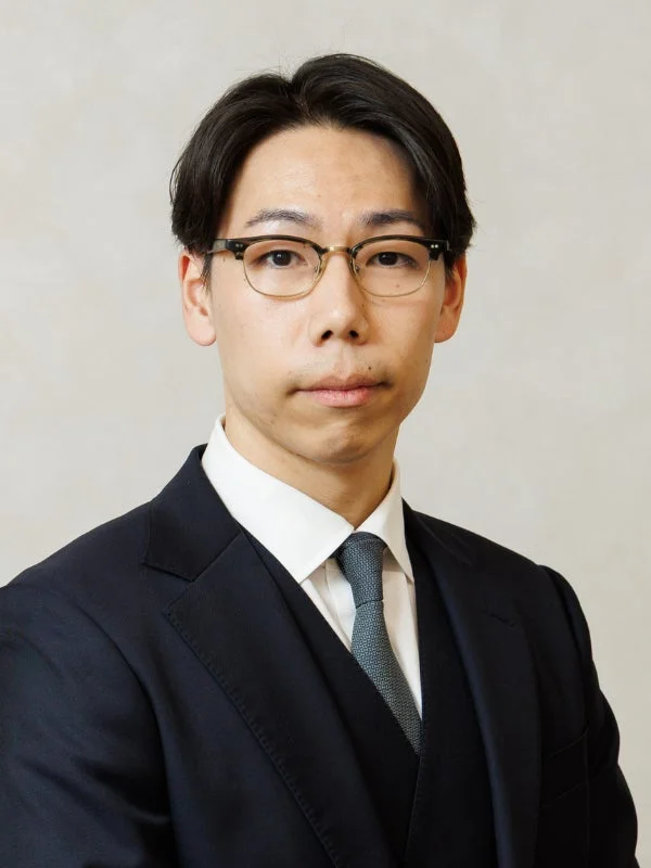 ダークスーツと眼鏡を着用した若いアジア人男性が、真剣な表情でカメラを見つめている上半身のポートレート写真です。ビジネスシーンに適した、清潔感のあるフォーマルな印象を与えます。