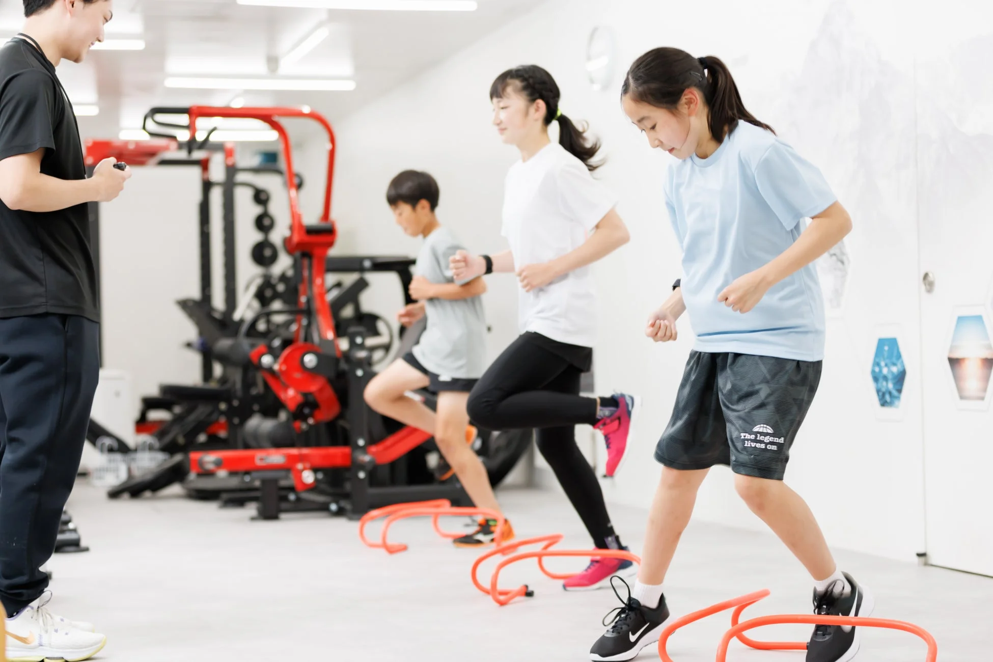 室内ジムで、コーチの指導のもと、子供たちがミニハードルを使ったアジリティトレーニングに励んでいます。