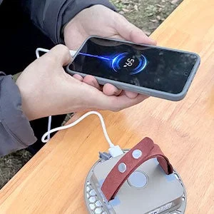 屋外の木製テーブルで、一人の人物がポータブル電源らしき機器を使ってスマートフォンを充電している様子です。スマートフォンの画面にはバッテリー残量「59%」が表示されています。