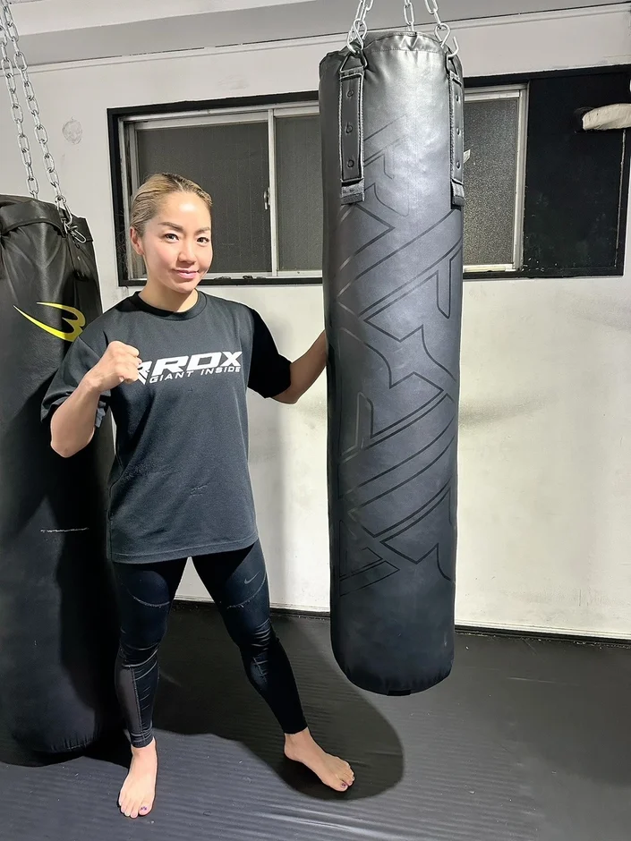 スポーツウェアを着た女性がジムでサンドバッグの横に立ってポーズをとっています