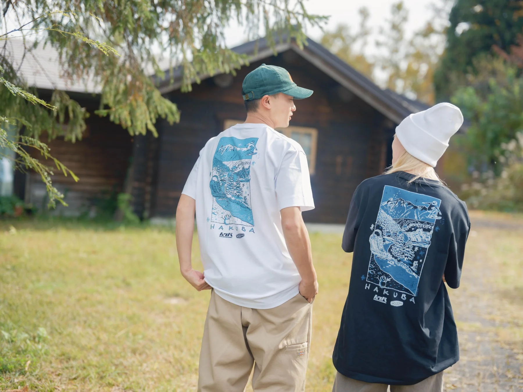 ログハウスの前で後ろ姿を見せる男女のカップル。白馬の冬景色が描かれたTシャツを着用