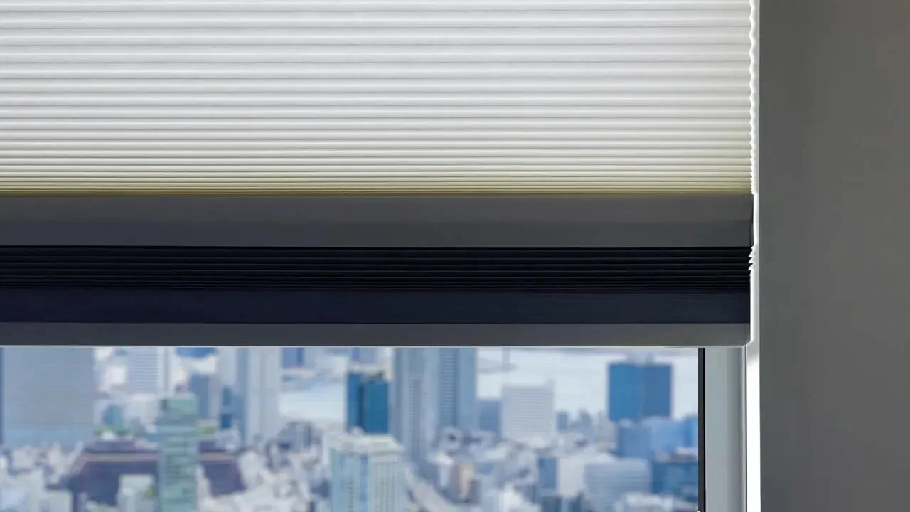 窓辺に設置された白と濃いグレーのツートンカラーのブラインドと、その下に見えるぼやけた都市の景色
