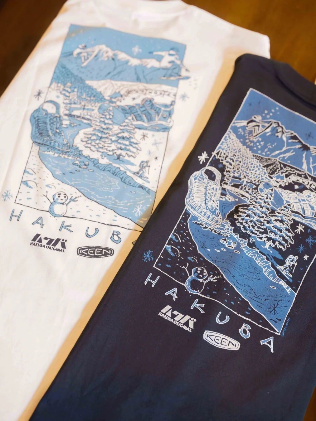 白と紺の2枚のTシャツが並べられ、雪山やスキーヤー、スノーボーダー、KEENのブーツなどが描かれた冬のアウトドアデザイン