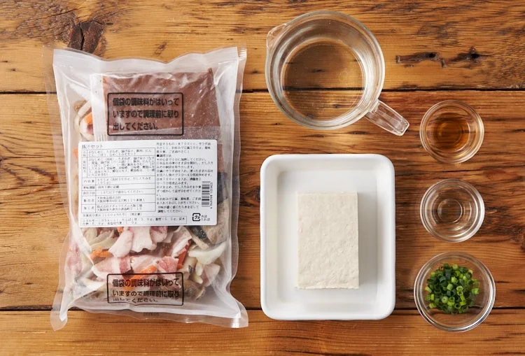 木製のテーブルに、豚肉や野菜、調味料が入った鍋セットの袋、豆腐、水、薬味などがきれいに並べられている様子
