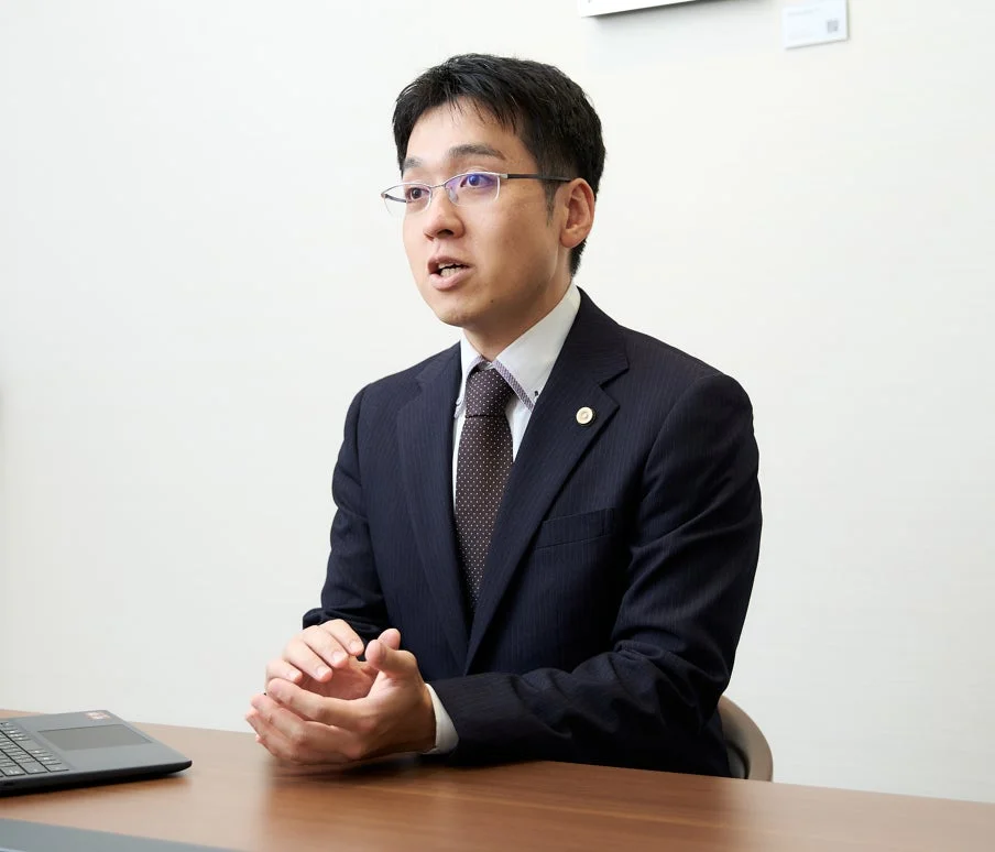 スーツを着て眼鏡をかけた男性が、木製の机に座り、身振り手振りを交えながら話している様子です。ビジネスシーンやインタビューの一場面と考えられます。