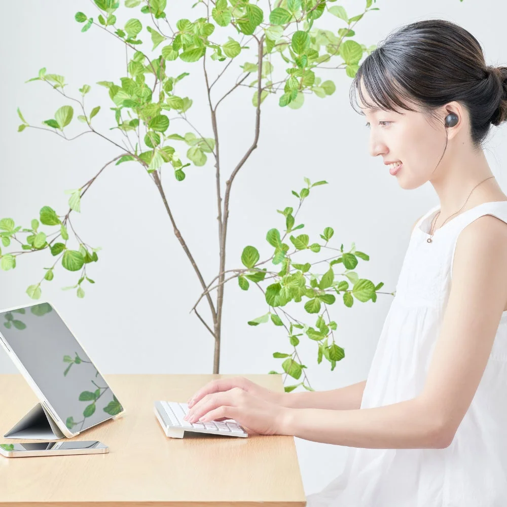 白い壁と緑の植物を背景に、白いワンピースを着た若い女性が木製の机で作業している様子。