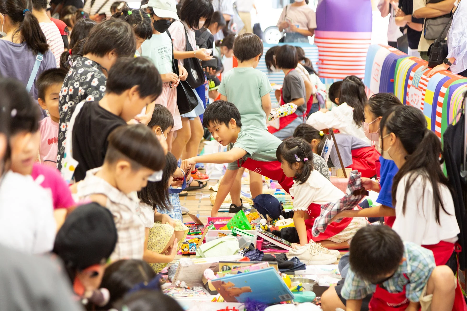 多くの子供たちが集まり、床に広げられた様々な商品やアイテムを熱心に見たり触ったりしています。賑やかな雰囲気のイベントや市場の様子です。