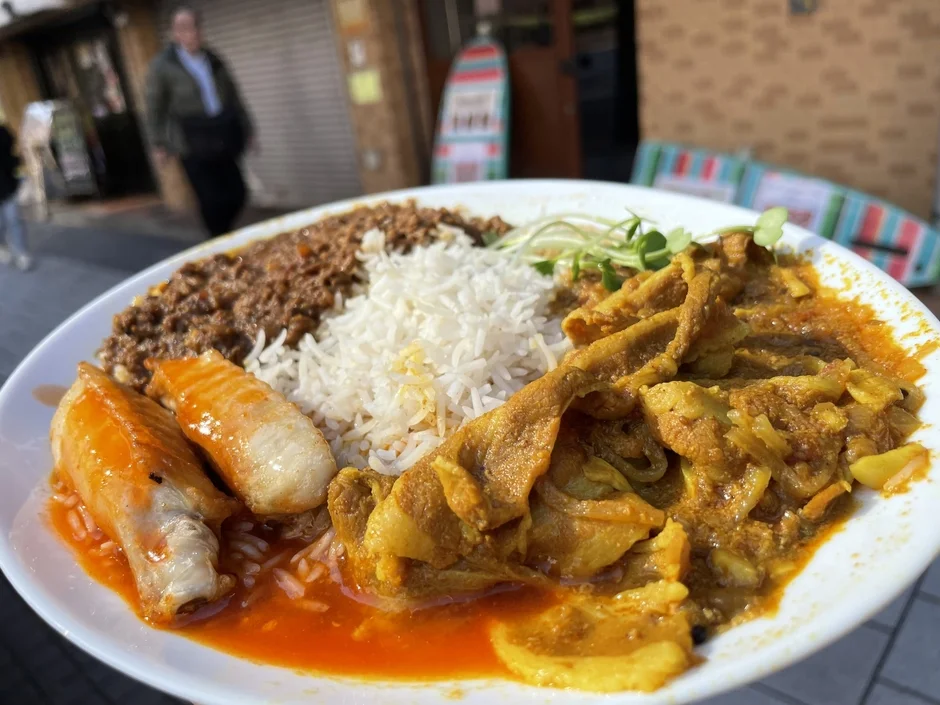 屋外で提供されているプレート形式の食事。白いご飯を中心に、ひき肉らしきもの、黄色いカレー風味の煮込み料理、赤いソースがかかった魚料理が盛り合わせになっている。カジュアルな雰囲気のランチまたはディナー。