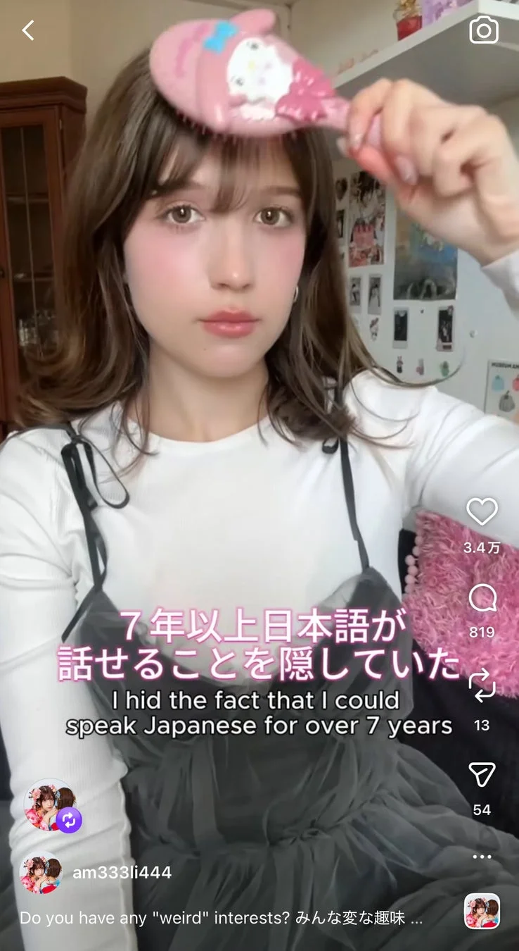 ピンクのマイメロディのヘアブラシを頭にかざす若い女性の動画コンテンツのスクリーンショット。女性はカメラを見つめ、「7年以上日本語が話せることを隠していた」というテキストが表示されており、自身の秘密を明かしている様子です。