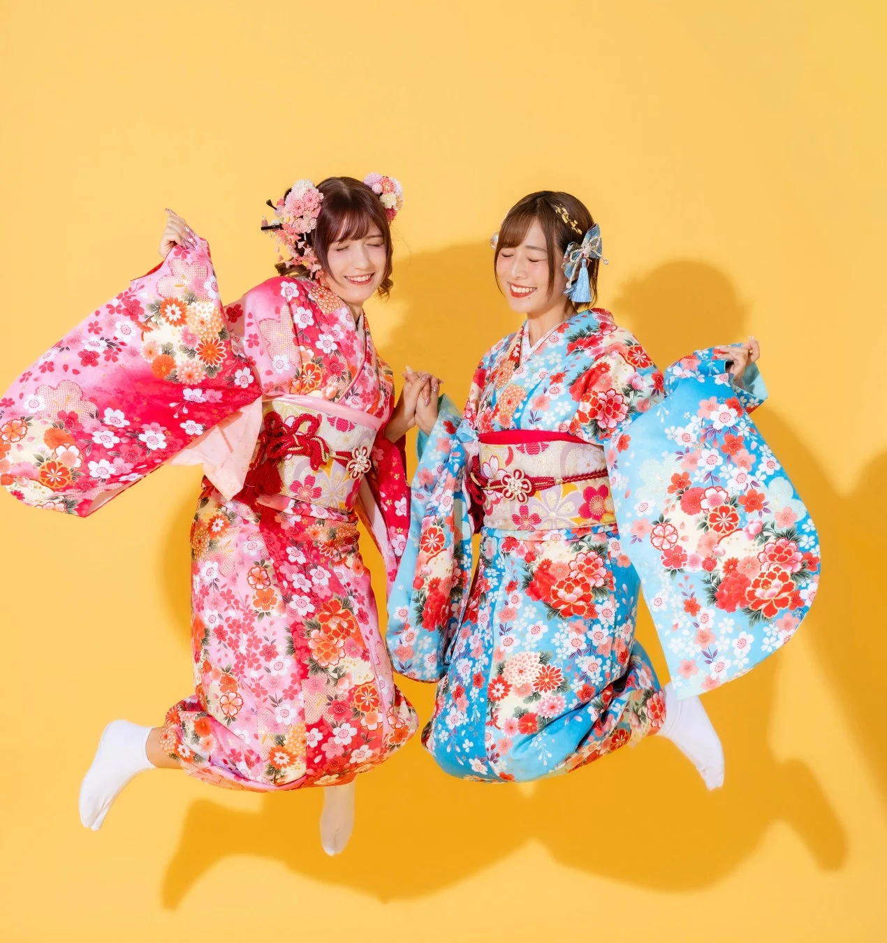 鮮やかな花柄の振袖を着た二人の女性が、手をつないで楽しそうにジャンプしている様子。笑顔が印象的で、成人式などのお祝いのシーンを連想させます。