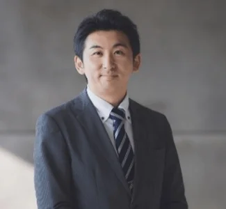 スーツを着用した若い男性が、カメラに向かって穏やかな笑顔を見せている上半身のポートレート写真