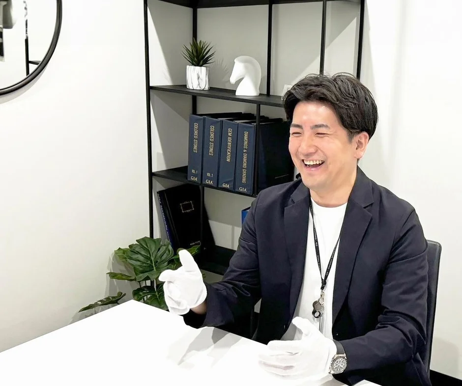 オフィスで白手袋をはめた男性が笑顔で座っています。背景の棚にはGIA関連の宝石鑑定書籍が並び、専門的な接客やコンサルティングの場面を写しています。