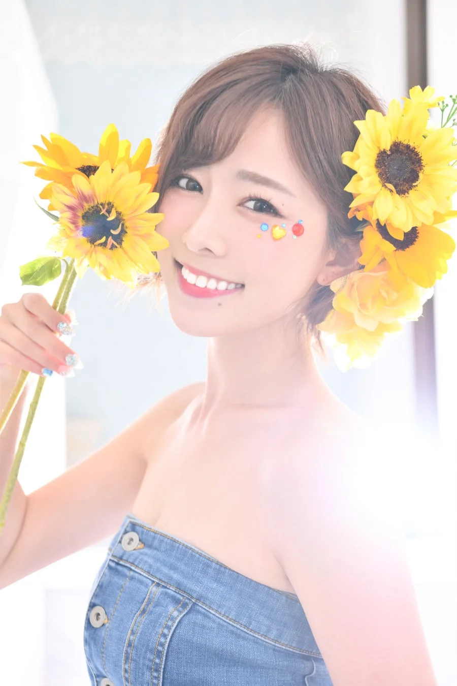 笑顔の女性がひまわりを手に持ち、肩出しのデニムトップスを着てポーズをとっている。頬にはキラキラした飾りが施され、髪にもひまわりが飾られており、夏の明るい雰囲気が伝わるポートレート写真です。