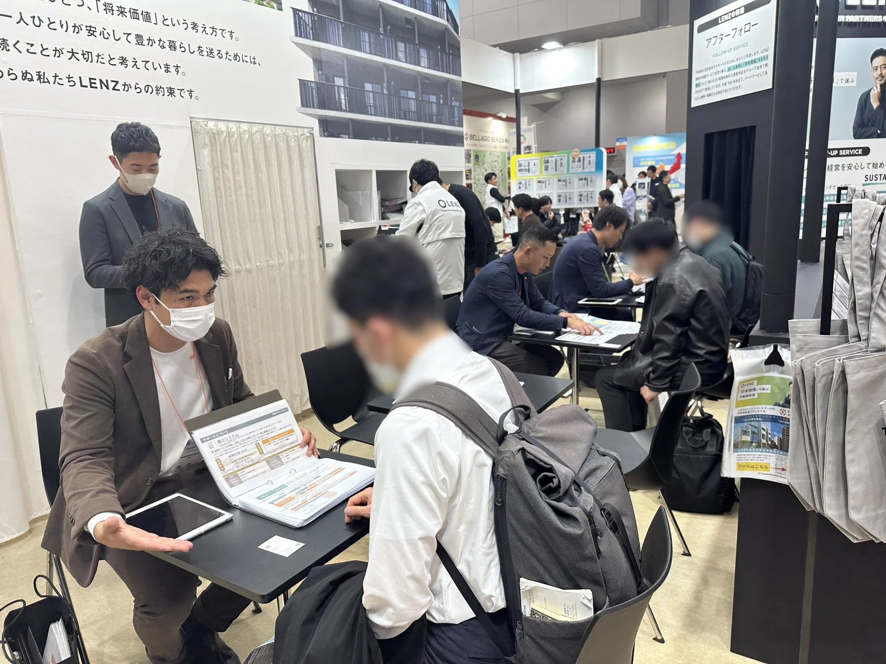 展示会またはセミナー会場で、男性が来場者に対し、資料やタブレットを使ってLENZのアフターフォローや不動産に関する説明を行っている様子