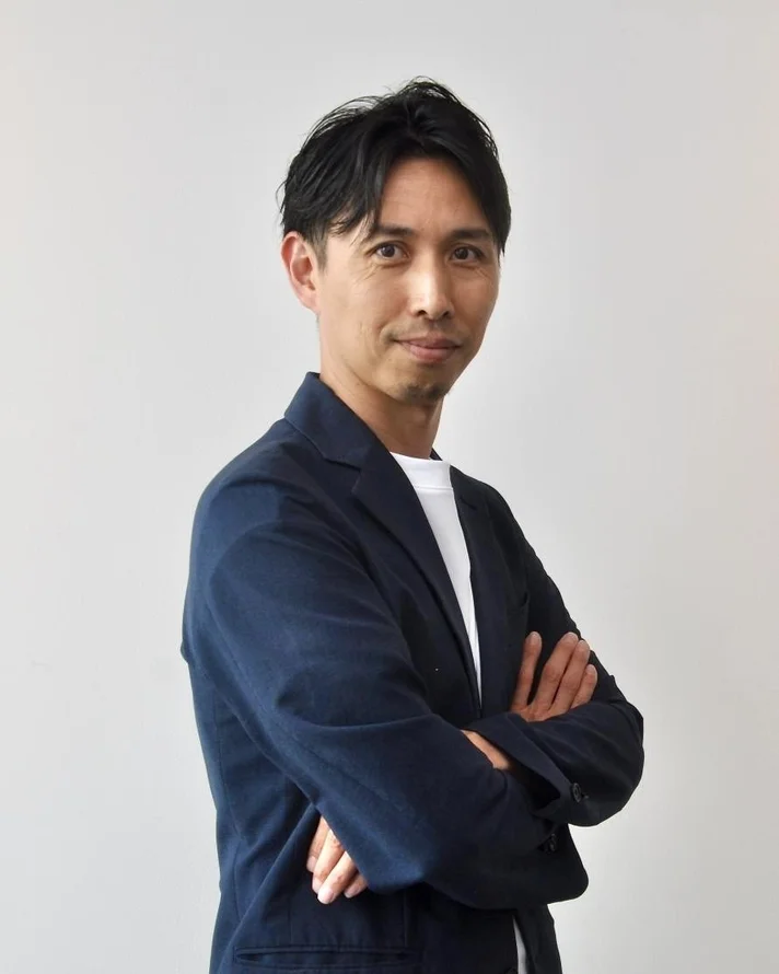 白いTシャツの上に紺色のブレザーを羽織った男性が、腕を組んでカメラを見つめているポートレート写真