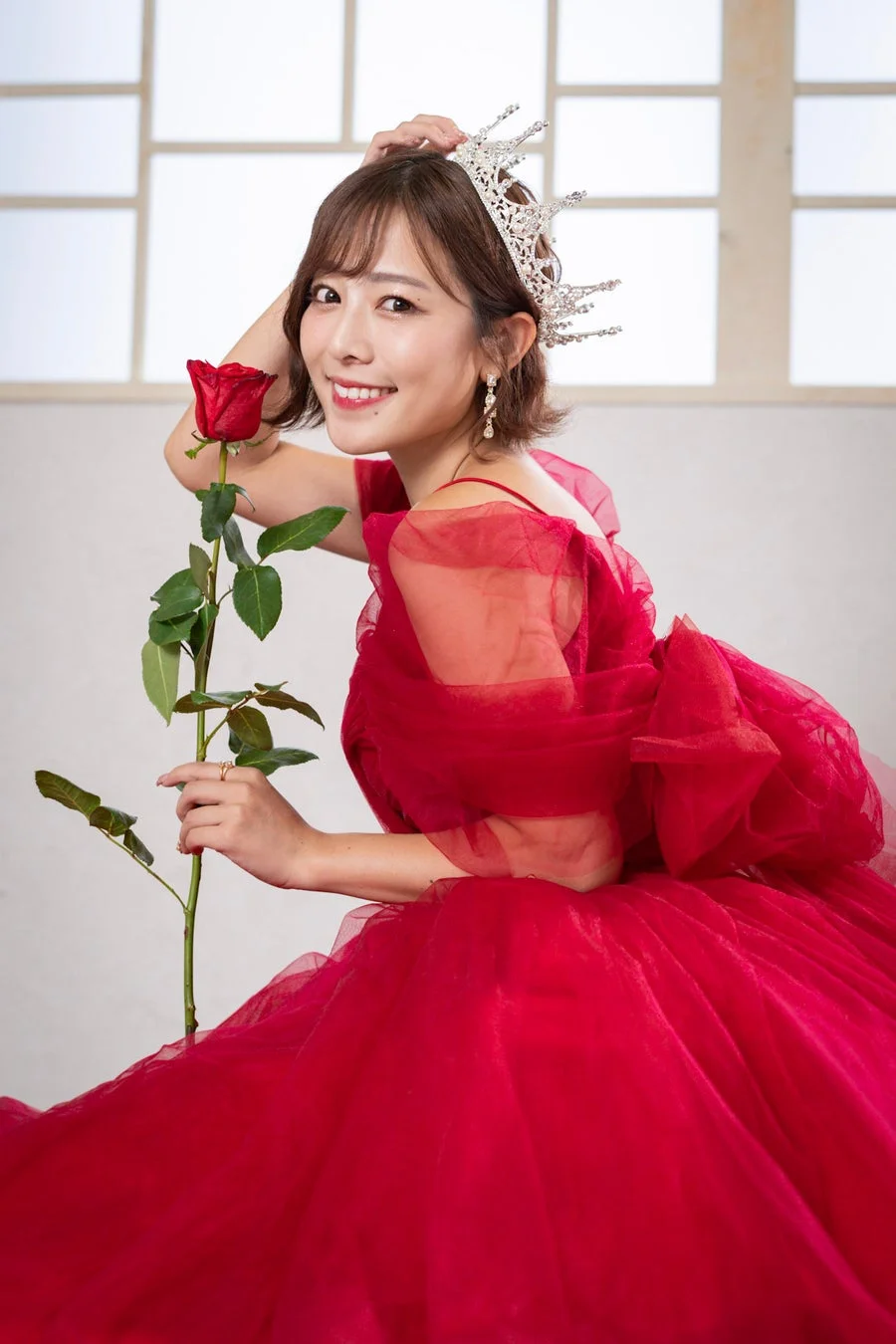 赤いドレスとティアラを身につけ、笑顔で赤いバラを持つ女性のポートレート。華やかで美しい印象の記念写真です。
