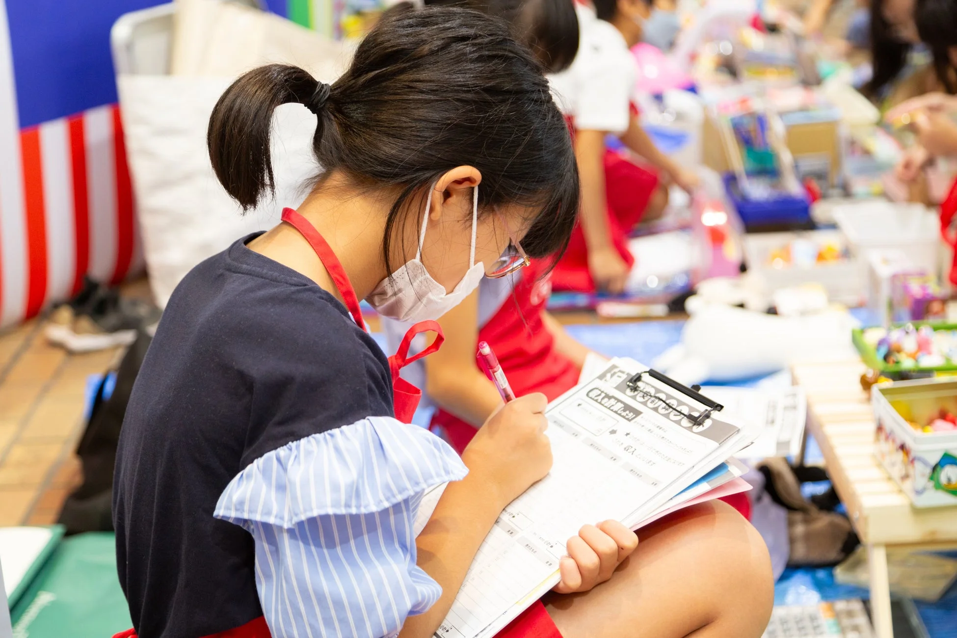 マスクとエプロンを着用した少女が、クリップボードに「収入を計算しよう！」と書かれた用紙にペンで書き込んでいる。学習イベントかワークショップに参加し、集中して作業に取り組む様子。