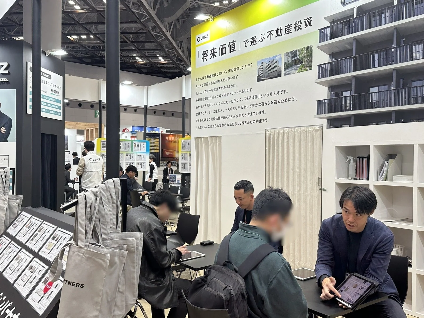 不動産投資に関する展示会ブースで、スタッフが来場者に対しタブレットを使って物件を説明している様子