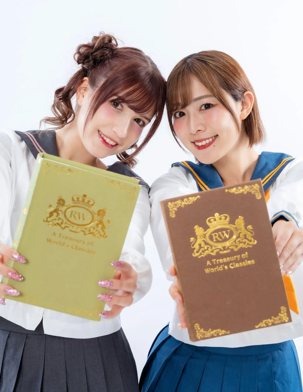 セーラー服を着た二人の女性が笑顔で本を掲げています。彼女たちは親密な雰囲気で、白い背景の前でポーズをとっています。