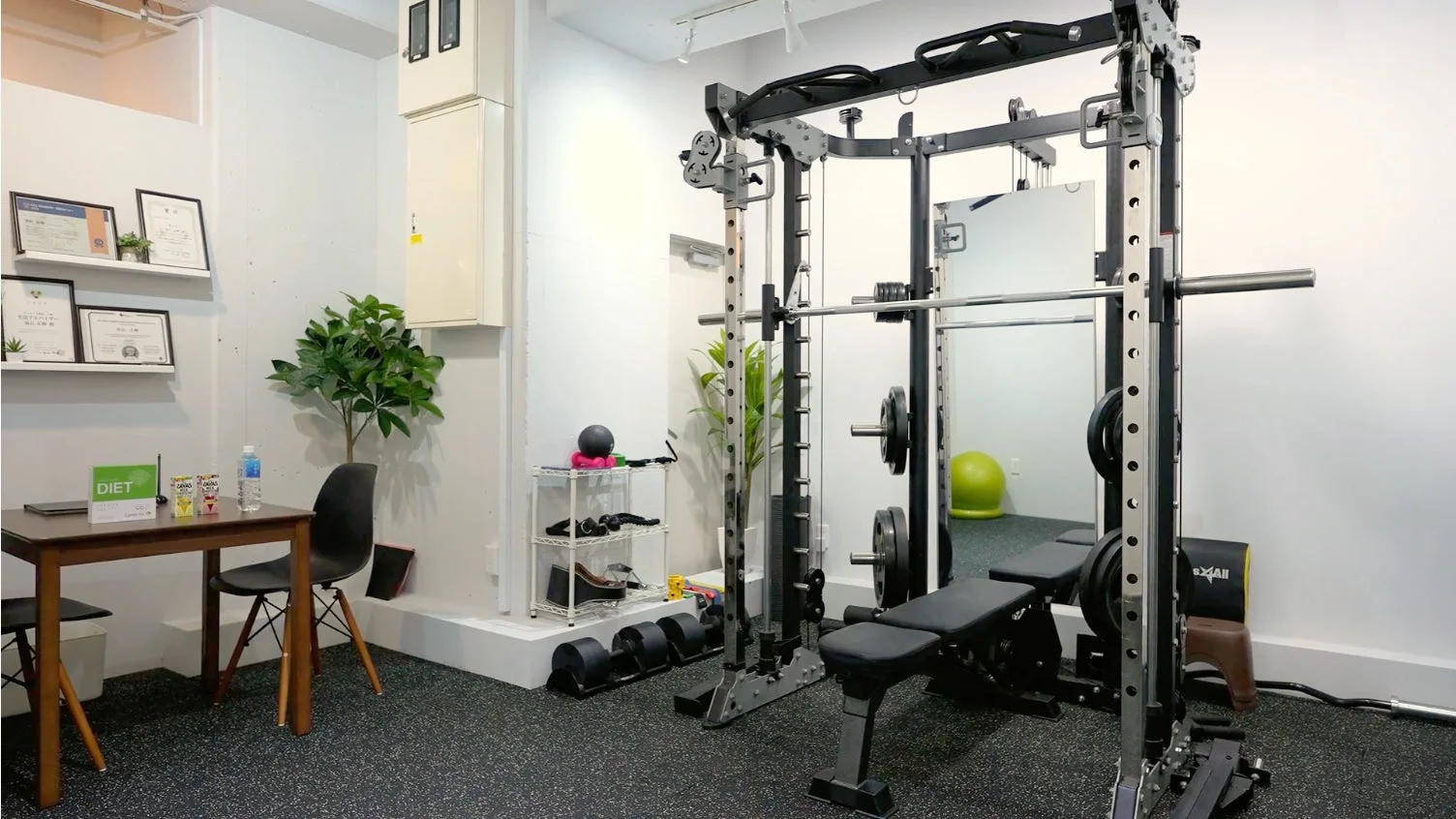 清潔感のある白い壁の室内ジムの様子です。最新のウェイトトレーニングマシン、ダンベル、ベンチが設置されており、パーソナルトレーニングに適した環境に見えます。奥にはカウンセリング用のテーブルと椅子、そして「DIET」と書かれた看板も見えます。