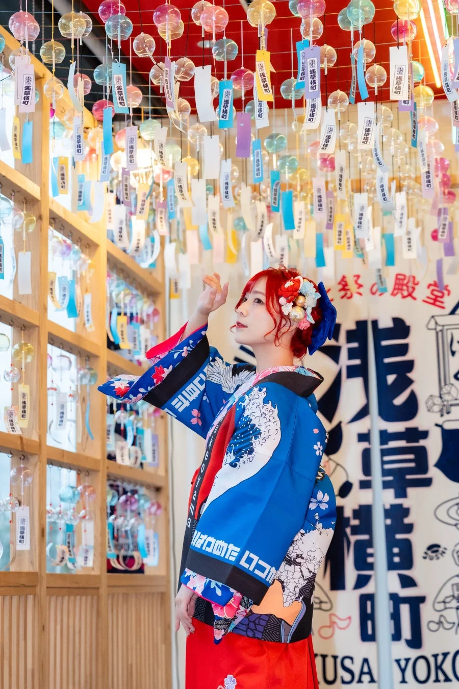 カラフルな風鈴が飾られた空間で、鮮やかな着物を着た赤髪の女性が上を見上げている。浅草横町の祭りの雰囲気が伝わる、和モダンなポートレート写真。