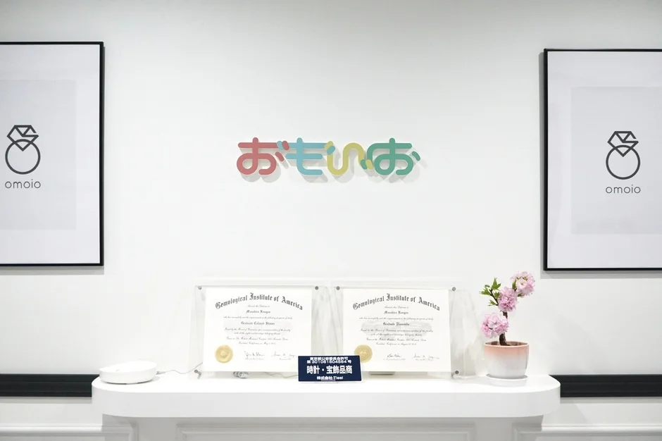 白い壁に「おもいお」のロゴとダイヤモンドリングの額縁が飾られ、棚にはGIA宝石鑑定士の資格証明書などが置かれた受付風景