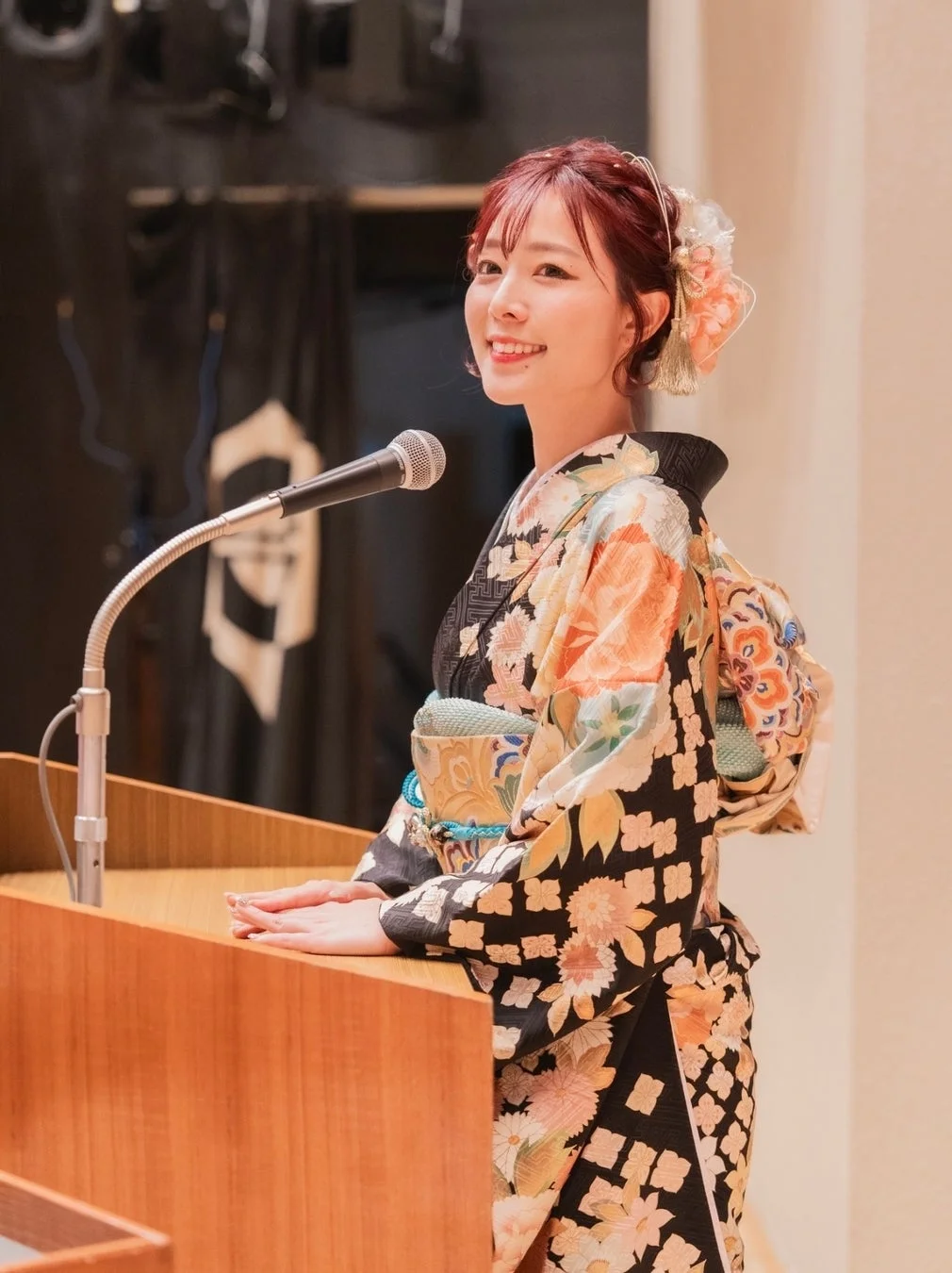 華やかな着物を着た女性が演台の前に立ち、マイクに向かって笑顔で話している様子です。