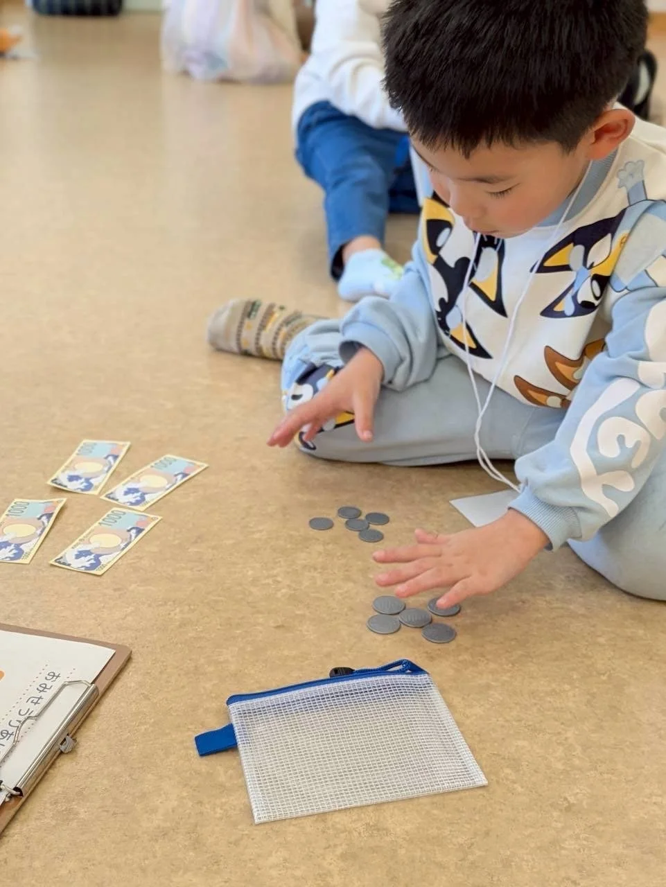 床に座った子供が、おもちゃの紙幣と硬貨を使ってお金の学習をしています。おつりの計算をしているようで、教育的な遊びの様子がうかがえます。
