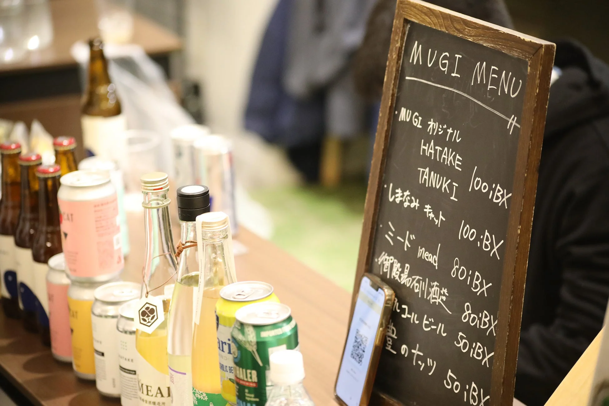 カウンターに並べられた様々なボトルや缶の飲み物と、手書きのMUGIメニューが書かれた黒板が写っています。