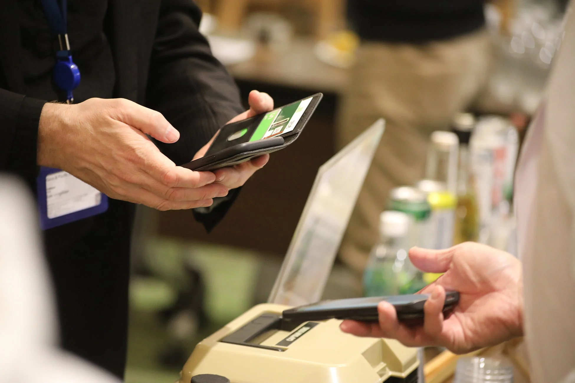 会場のような場所で、2人の人物がスマートフォンやカードを使って何らかの登録や支払いのやり取りをしている様子。