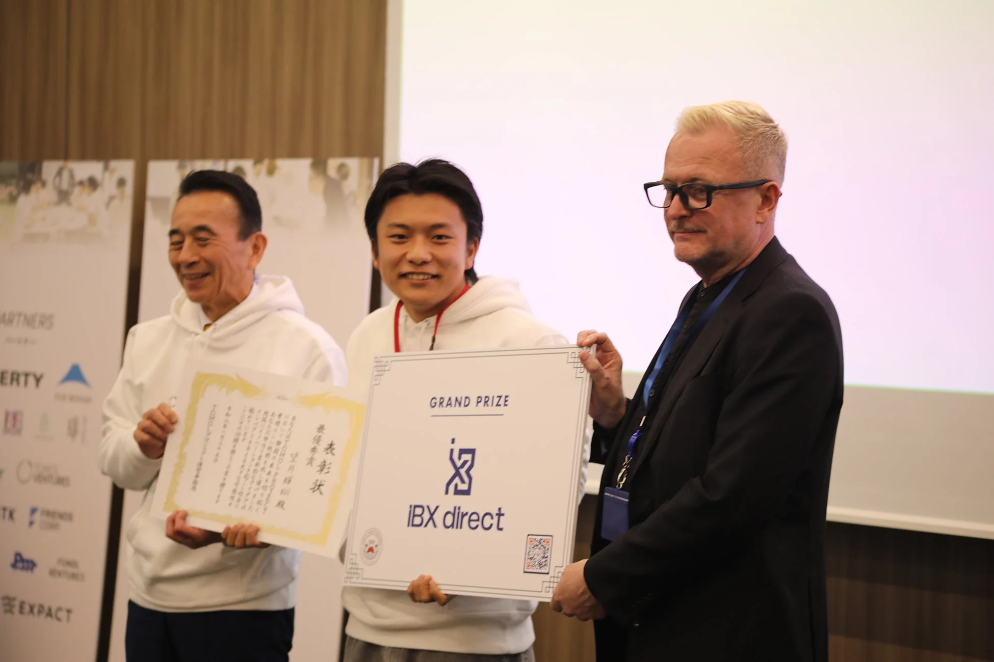 ビジネス関連の授賞式で、白いパーカーを着た受賞者とスーツ姿のプレゼンターを含む男性3名が、表彰状と「GRAND PRIZE IBX direct」のボードを掲げている。