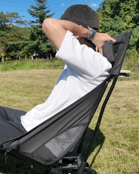 屋外で男性が黒いポータブルチェアに座り、両手を頭の後ろに組んでリラックスしている様子