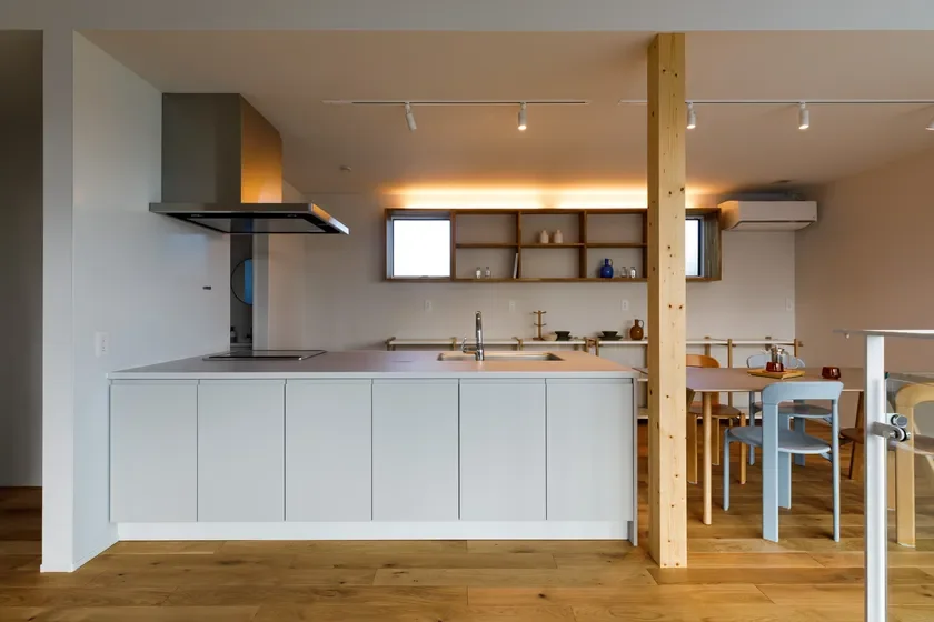 ライトグレーのアイランドキッチンと木製シェルフが特徴的なモダンなLDK。温かい照明とフローリングが心地よい空間を演出しています。