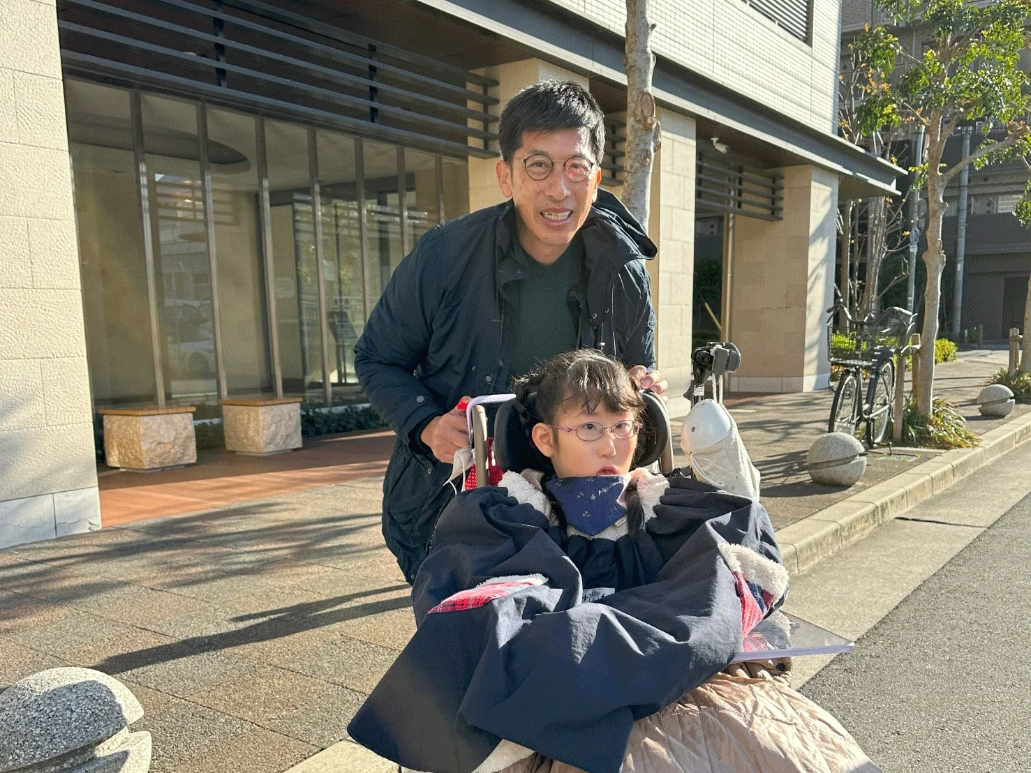 晴れた日に、男性が車椅子に乗った少女を屋外で押している様子