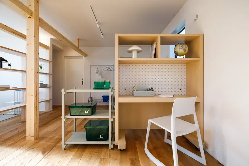 明るい木目の床と白い壁が特徴のモダンなインテリア空間。木製のデスクと収納棚があり、白い椅子や緑色の収納ボックス、地球儀などが配置されています。シンプルで機能的な書斎またはリビングの一角を捉えた写真です。