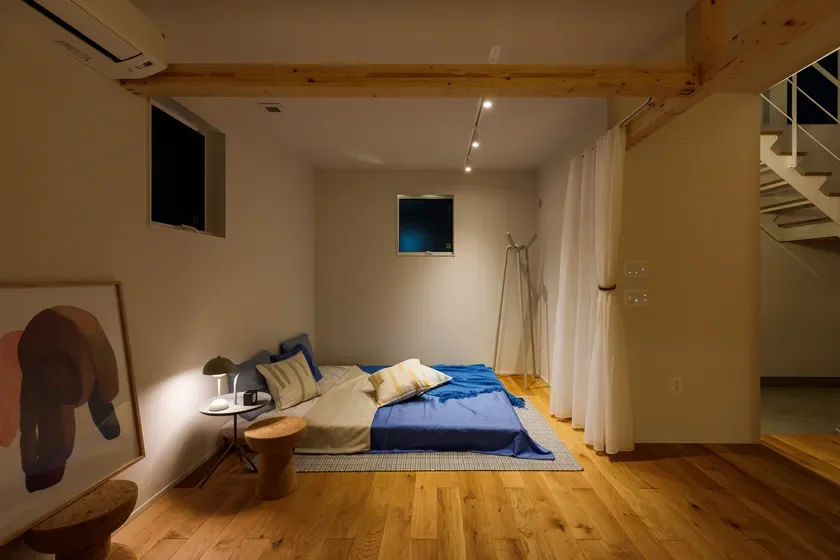 木製フローリングと天井の梁が特徴のモダンな寝室。ブルーとベージュの寝具が置かれたローベッドと、温かい間接照明が落ち着いた空間を演出。抽象画がアクセントになっています。