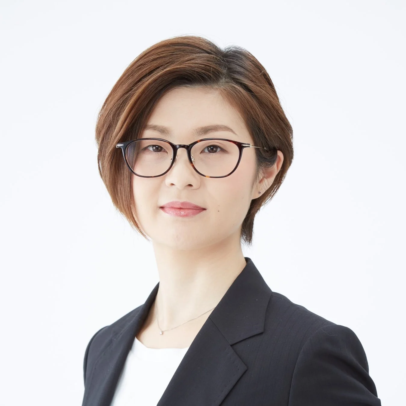 白い背景の前で、眼鏡をかけたショートヘアの女性が、黒いジャケットを着用し、カメラをまっすぐ見つめているビジネスポートレート