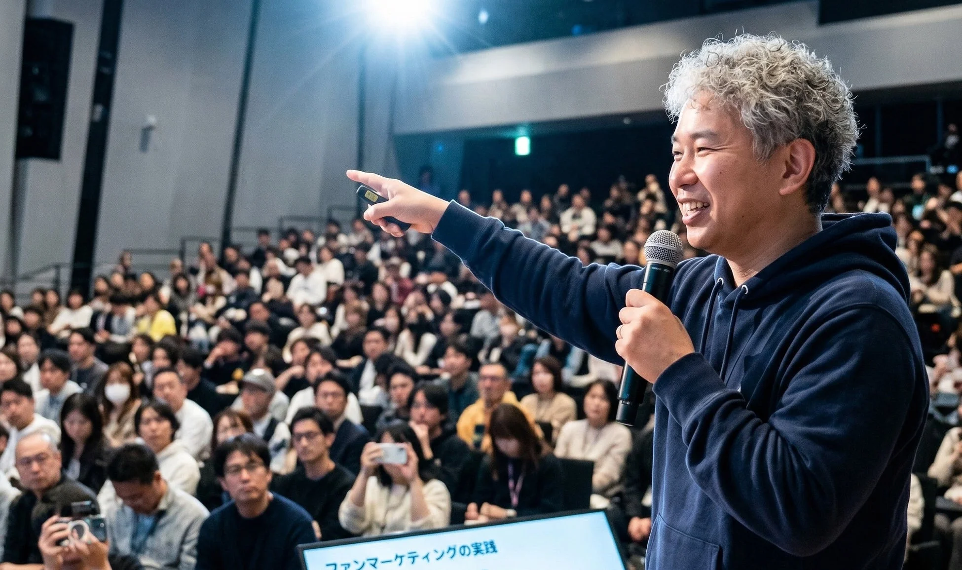 講演会場で、笑顔の男性がマイクを持ち、大勢の聴衆に向けてプレゼンテーションを行っています。スクリーンには「ファンマーケティングの実践」と表示されています。